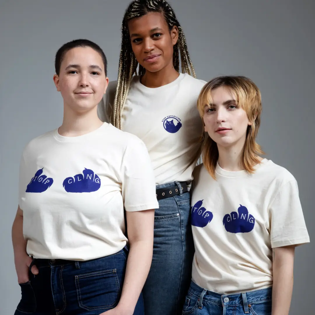 Drei Personen in weißen T-Shirts mit dunkelblauen Aufdrucken: Zwei tragen Schriftzüge über Katzenpfoten mit ‚STOP‘ und ‚CLNG‘, eine trägt das ‚Stop Catcalling‘-Logo.