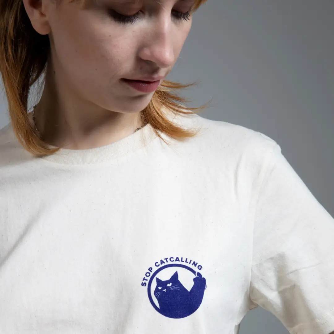 Person in weißem T-Shirt mit blauem Logo, das eine Katze zeigt und den Schriftzug ‚Stop Catcalling'