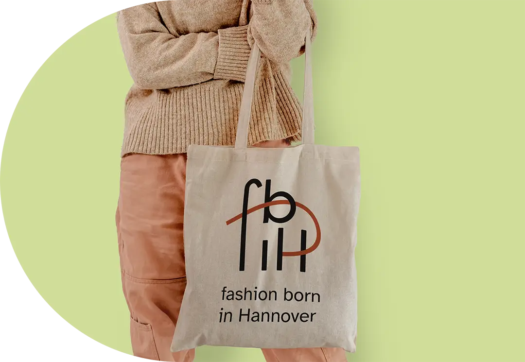 Unterk&ouml;rper einer Person in schlichter Kleidung, die einen hellbraunen Jutebeutel h&auml;lt. Auf diesem ist das fashion born in Hannover - Logo zu sehen.
