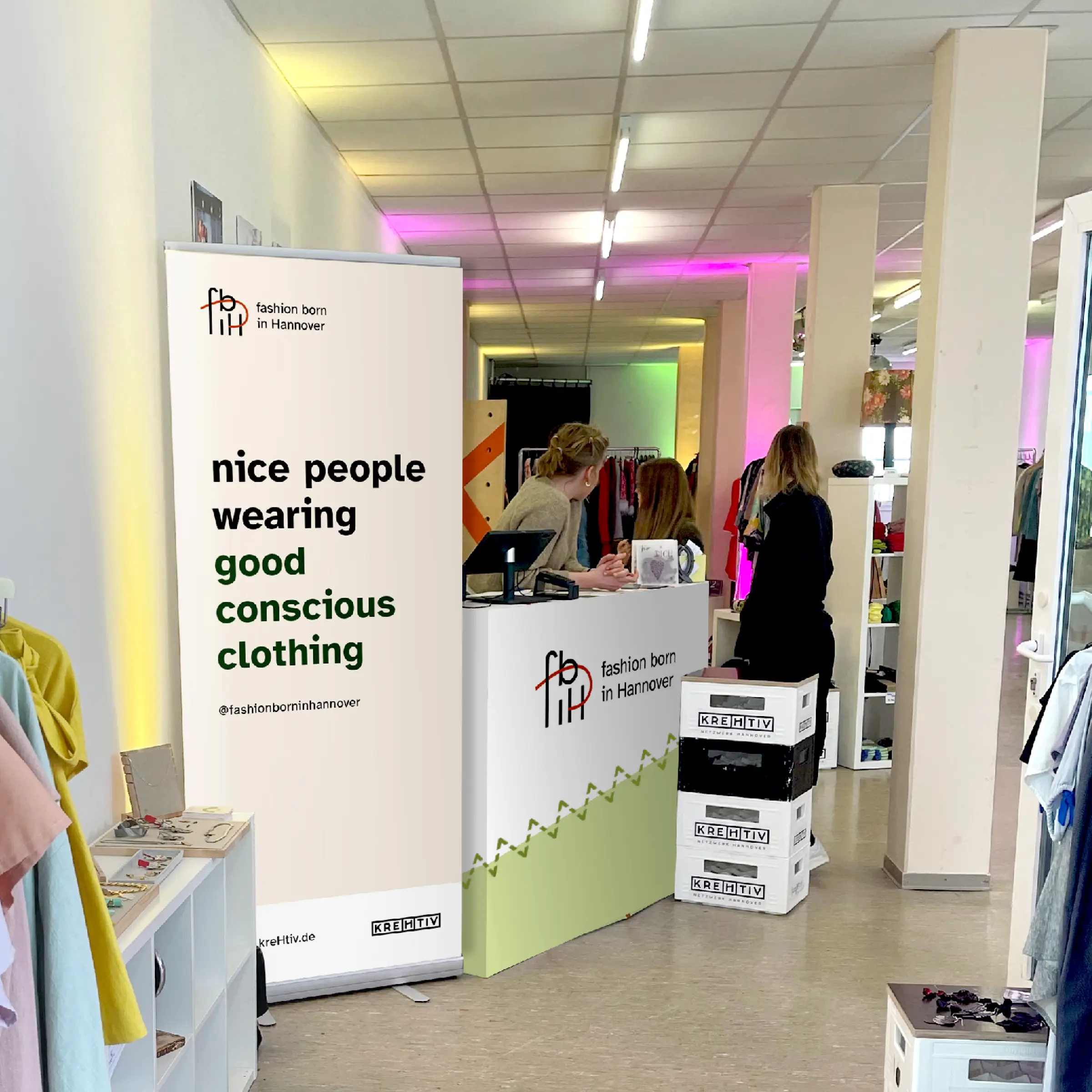 Das Foto zeigt einen hellen Verkaufsraum mit Menschen, die vertreut im Raum stehen. Im Fokus steht ein hochkantiges Roll-Up-Banner, welches ein helles Layout mit der Aufschrift: nice people wearing good conscious clothing, zeigt.