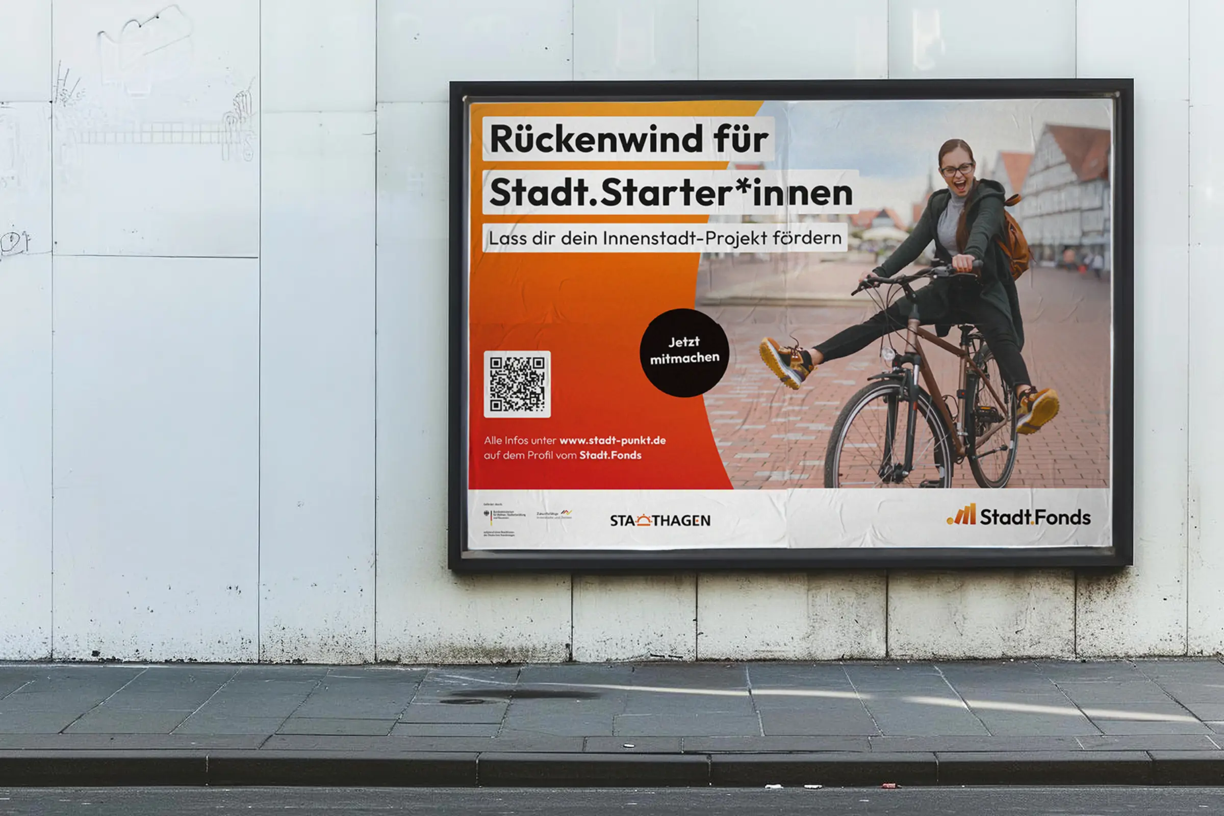 Darstellung eines querformatigem OOH-Plakats an einer Au&szlig;enwand. Das Plakat zeigt eine Frau, die energetisch auf einem Fahrrad sitzt und in die Kamera schaut. Daneben steht eine Headline: R&uuml;ckenwind f&uuml;r Stadt.Starter*innen und kommuniziert die Inhalte der Submarke Stadt.Fonds.
